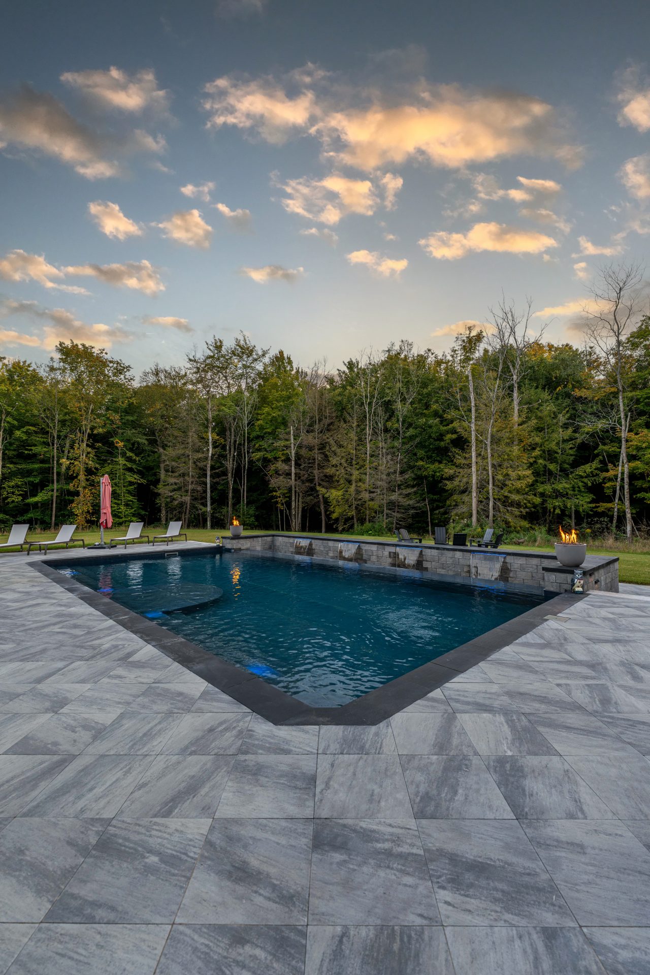 Rectangular pool with fire pits and forest backdrop.