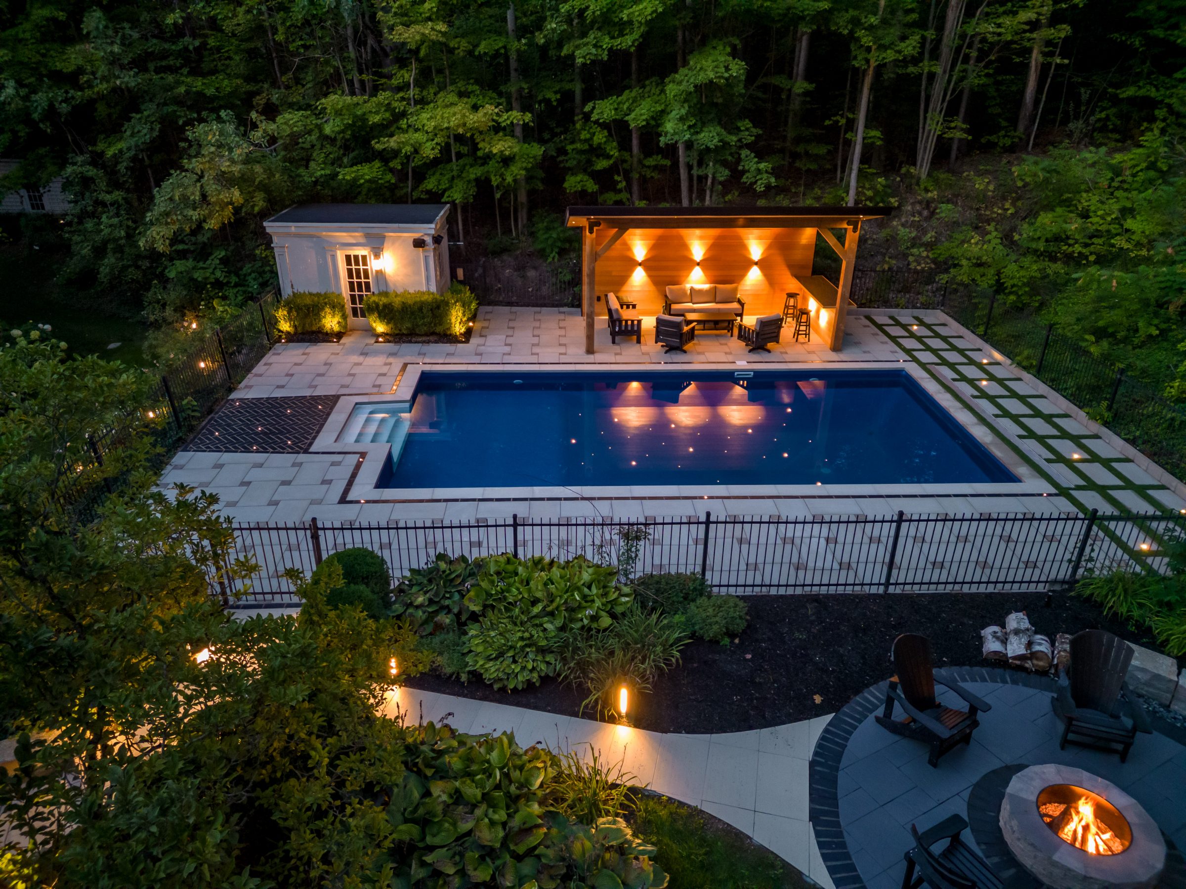 Illuminated backyard pool with seating and fire pit.