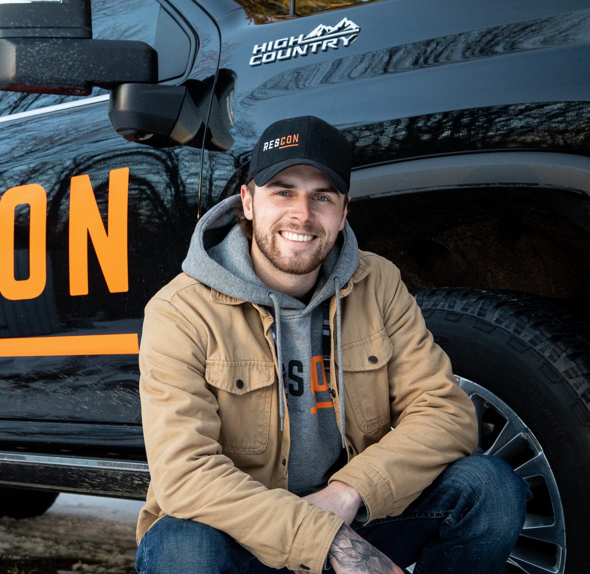 Smiling man crouching beside black truck. Outdoor vibe.